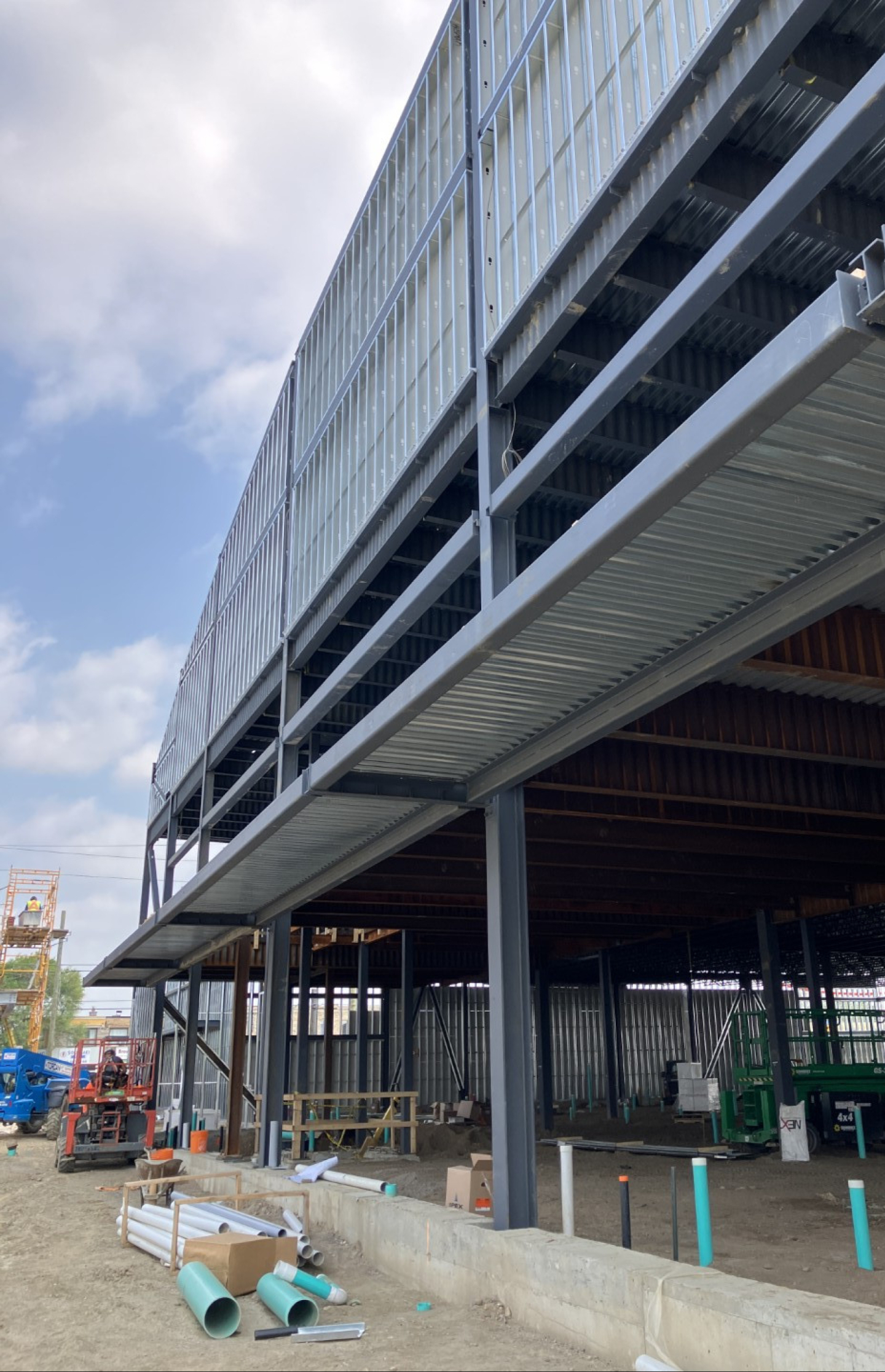 An unfinished industrial building exterior with exposed steel framing and metal wall panels. Construction materials, utility pipes, and equipment sit along a concrete foundation beneath a partly cloudy sky.